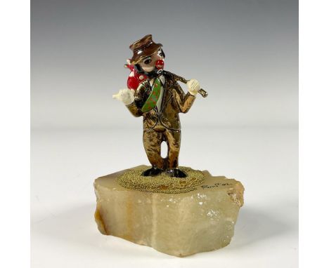 A gilded enamel figure of a smiling clown wearing a green jacket, large red bow tie, and blue pants. Presented on a white ony