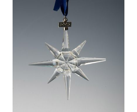A clear, faceted eight point snowflake adornment featuring a metal tag with 1995 impressed on it, hanging from a navy blue ri