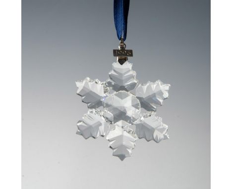 A clear, faceted six point leaf-like snowflake adornment featuring a metal tag with 1996 impressed on it, hanging from a sati