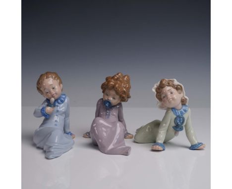 A delightful trio of Lladro porcelain figurines depicting young children seated in relaxed, sleepy poses, each dressed in pas