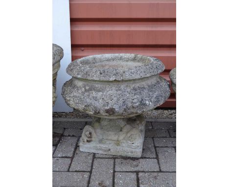 A large weathered reconstituted stone mortar-shaped planter,  standing on a separate plinth with dolphin masks to the corners