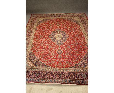 A KASHAN CARPET, the finely woven claret red field with navy blue spandrels and central gul and all over scrolling floral pat