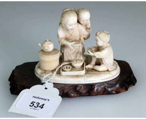 A Meiji Period carved ivory okimono, of a mother with child on her back offering a bowl of rice to a boy seated before a low 