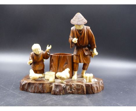 A JAPANESE WOOD AND IVORY GROUP OF A FATHER AND CHILD EITHER SIDE OF A TREE TRUNK AMONGST CHICKENS.   W.20.5CMS.