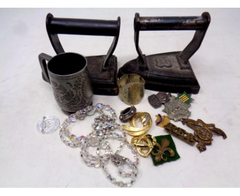 A box of two antique irons, plated napkin ring, pewter tankard, costume jewellery, military badges, Northumberland silver ena