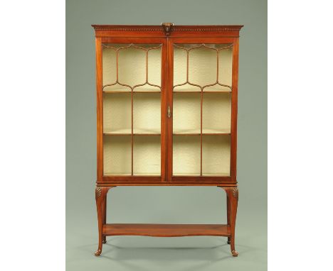 An Edwardian mahogany display cabinet, with mask and dentil cornice above a pair of glazed doors enclosing shelves and all ra