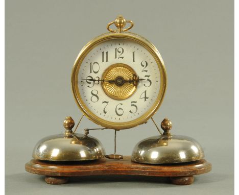 An Edwardian alarm clock, the brass case with suspension loop surmounted above two plated bells, all upon a shaped wooden bas