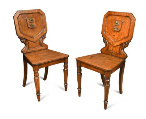 A pair of William IV oak hall chairs, with applied brass armorial crests to the panel backs with leaf carved detail, on octag