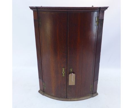 A Georgian bow fronted oak corner cabinet having a dentil cornice over two cupboard doors, flanked by fluted detail, H 94 x W