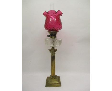 A Victorian brass oil lamp with a funnel, a cranberry glass shade and clear reservoir with line cut decoration, over a Corint