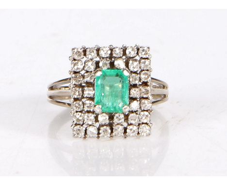 An 18 carat white gold emerald and diamond ring, having an emerald to the centre, set on a raised diamond set stepped plaque.