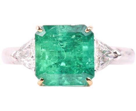 A vivid emerald and diamond three-stone ring, set with a central emerald measuring 9.2mm x 8.9mm, between trillion cut diamon