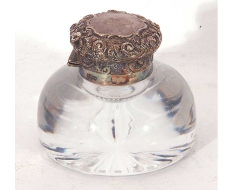 Circular clear glass inkwell with star cut base, hinged silver lid with embossed floral and foliate detail, a central cartouc