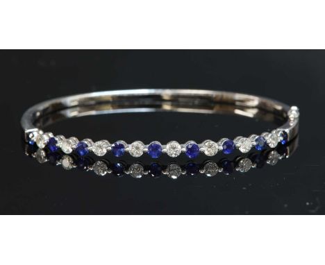 A white gold sapphire and diamond hinged bangle, with a row of brilliant cut diamonds and circular mixed cut sapphires, end s