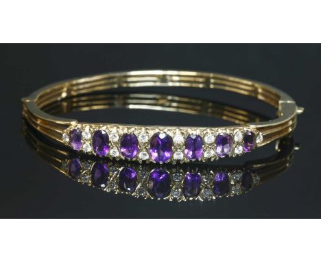 A 9ct gold amethyst and diamond hinged bangle, with a graduated row of oval mixed cut amethysts, claw set between pairs of br