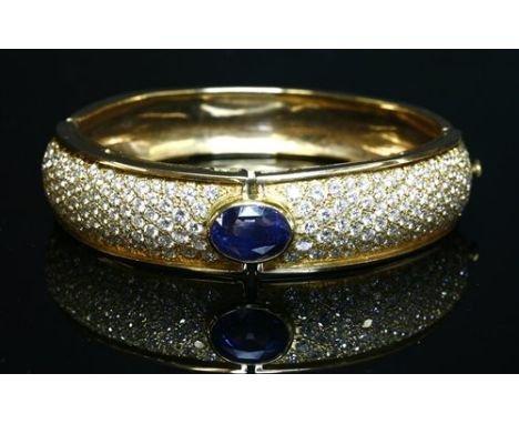 A gold, sapphire and diamond hinged bangle, with an oval mixed cut sapphire, rub set to the centre of a tapered top half, wit
