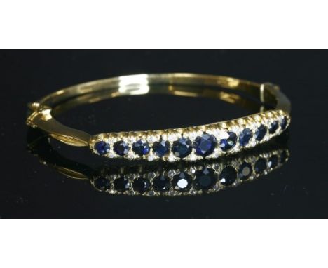 An 18ct gold, sapphire and diamond hinged bangle, with a row of circular mixed cut sapphires, with pairs of eight cut diamond
