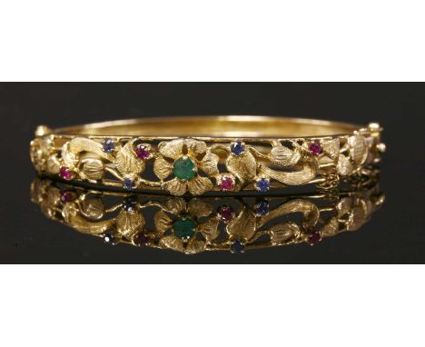 A 9ct gold emerald, ruby and sapphire set hinged bangle, c.1970, with a pierced foliate top half. A circular mixed cut emeral