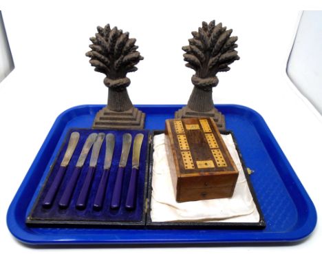 A tray of 19th century cast iron wheat sheaf door stops together with a set of cased butter knives and wooden cribbage board 