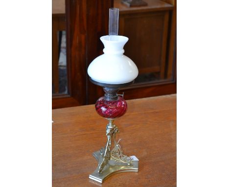 An oil lamp with cranberry glass reservoir and opaque glass shade, gilt metal column, pierced and decorated with rams heads, 