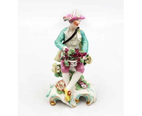 A Derby figure of a &lsquo;grape seller&rsquo; seated on a tree stump holding a basket of grapes in his lap. Circa 1760. Size