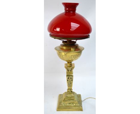 An ornate brass oil lamp with red glass shade, converted to electricity, height 67cm.