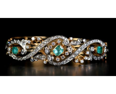 AN EMERALD AND DIAMOND STIFF HINGED BANGLE, the three step-cut emeralds collet set within a pierced and scrolled front set wi