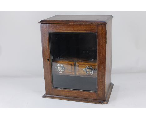 An oak smokers table top cabinet with glazed door enclosing pipe racks and two drawers, on plinth base (a/f) 28cm