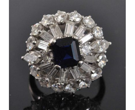 A white metal, sapphire and diamond bombe cluster ring, featuring a centre step cut sapphire within a border of twelve baguet