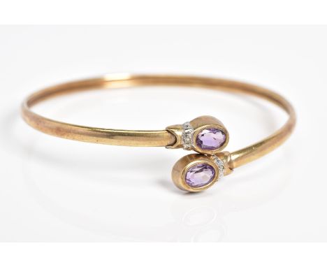 A 9CT GOLD CROSSOVER BANGLE, the open bangle set with two oval cut amethyst and single cut diamond detail, to the plain polis