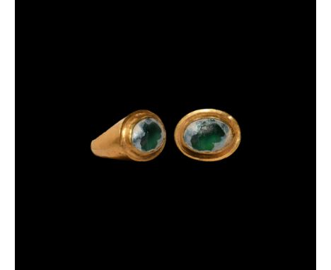 2nd century BC. A hollow-formed gold finger ring with inset green glass cabochon to the bezel, remains of iridescent surface 