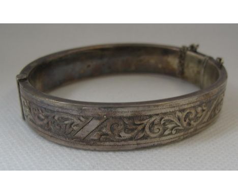 A silver bangle.  Hinged oval bangle decorated with engraved scrolling foliage.  With safety chain.(B.P. 21% + VAT) 