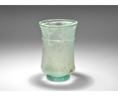 3rd-5th century AD. A pale blue glass cup of waisted profile with integral foot, chamfered rim, applied trail to the waist. C
