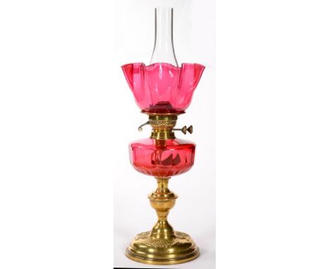 A LATE VICTORIAN EMBOSSED BRASS OIL LAMP, WITH FACETED CRANBERRY GLASS FOUNT AND DUPLEX BURNER, WITH A FRILLED CRANBERRY GLAS