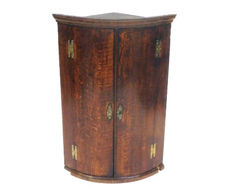 A George III oak bow fronted corner cupboard, with an outswept pediment over a pair of doors enclosing three shelves, on a pl
