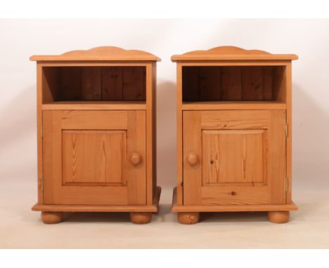 A pair of pine bedside cabinets - with a shaped back above an open shelf and a panelled cupboard door, raised on bun feet. (L