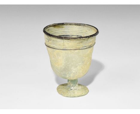 6th century AD. A pale blue glass cup with short stem and broad base; the bowl with black glass applied trails to the upper b