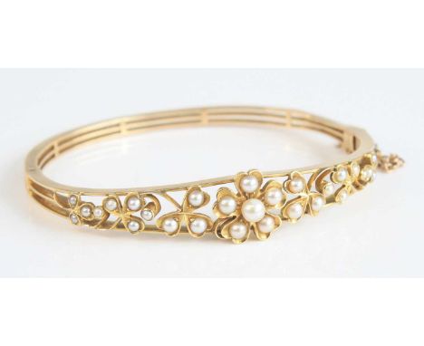 A yellow metal and seed pearl set hinged bangle, with safety chain, the top half set with a centre flower head and further tr