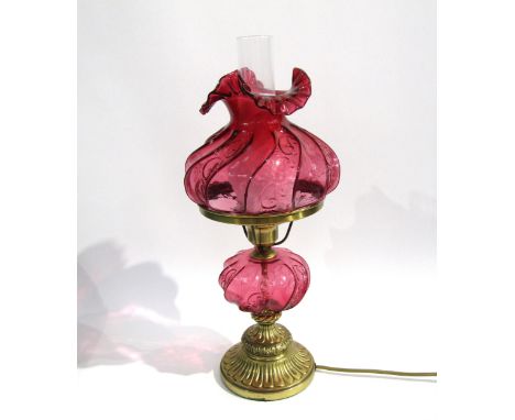 A cranberry glass table lamp with wrythen and frilled shade with polished brass support over glass reservoir with heavy cast 
