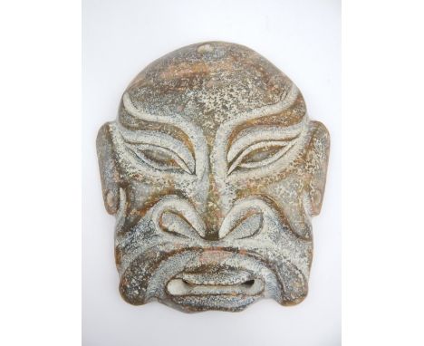 A large carved boulder jade Chinese mask, the stylised features set in a theatrical grimace, the opaque jade is speckled gree