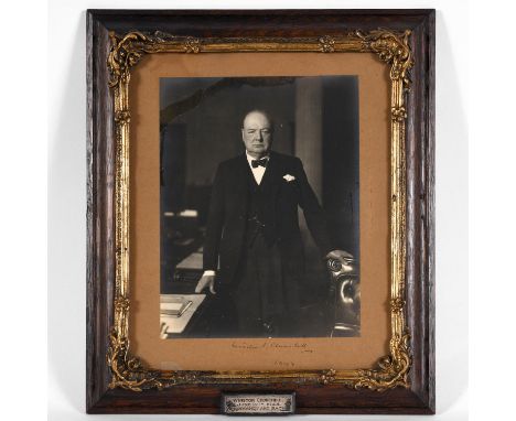 A large three quarter length photograph of Winston Churchill beside his desk, signed to the mount 'Winston Churchill, 1944'. 