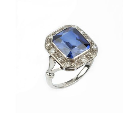 A SYNTHETIC SAPPHIRE AND DIAMOND CLUSTER RING, the octagonal mixed-cut synthetic sapphire collet set within a border of singl