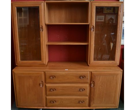 A light Ercol display cabinet, the top with a pair of glazed doors above a projecting base with three long drawers flanked by