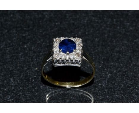 A sapphire and diamond cluster ring, central round cut blue sapphire, surround by a square platform encrusted with twelve rou