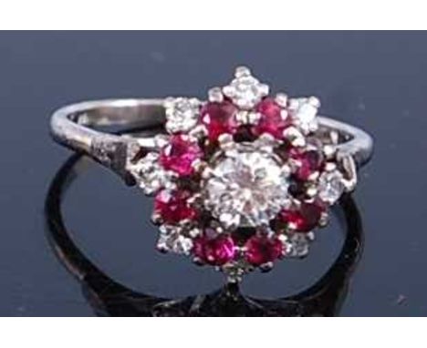 A white metal, ruby and diamond circular reverse cluster ring, featuring a centre round brilliant cut diamond within a border