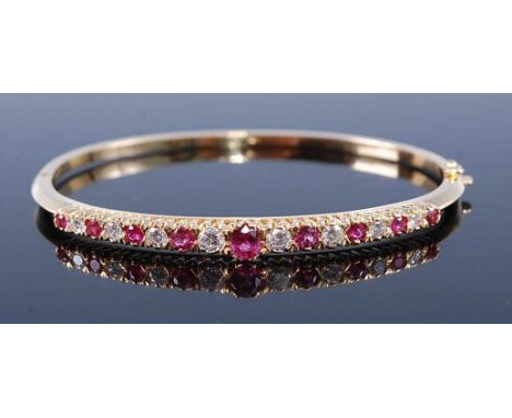 A yellow metal, ruby and diamond oval hinged bangle, having nine graduated rubies alternating with eight round brilliant cut 