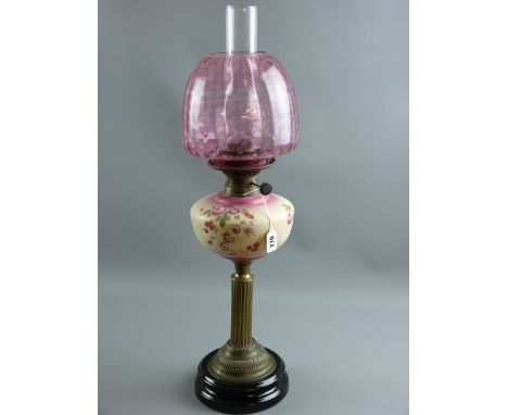 A Victorian oil lamp with floral decorated pottery font on a brass column and black pottery base with cranberry tinted shade 