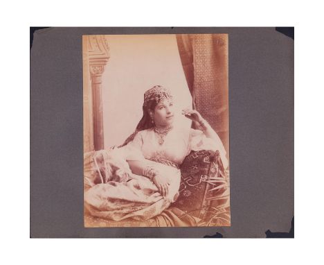 Sepia toned portrait of a seated Algerian woman in traditional dress and crown surrounded by tapestries and pillows. Fixed to