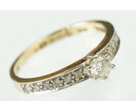 9ct gold and diamond ring. The ring being set with a round brilliant cut diamond to the head flanked by twelve round cut diam