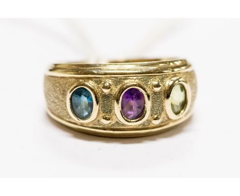 A three stone set ring, set with oval cut amethyst, blue topaz and peridot cabochons, rubover set to a textured band with pol